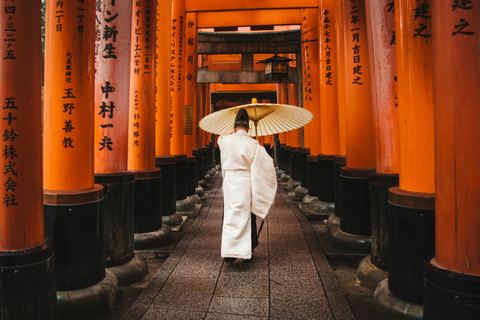 A woman in a white kimono with a paper umbrella walking through the orange torii gates of Fushimi Inari in Kyoto