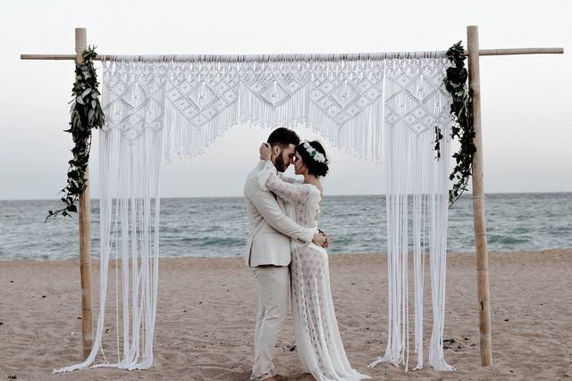A couple embracing under a macramé wedding arch on the beach